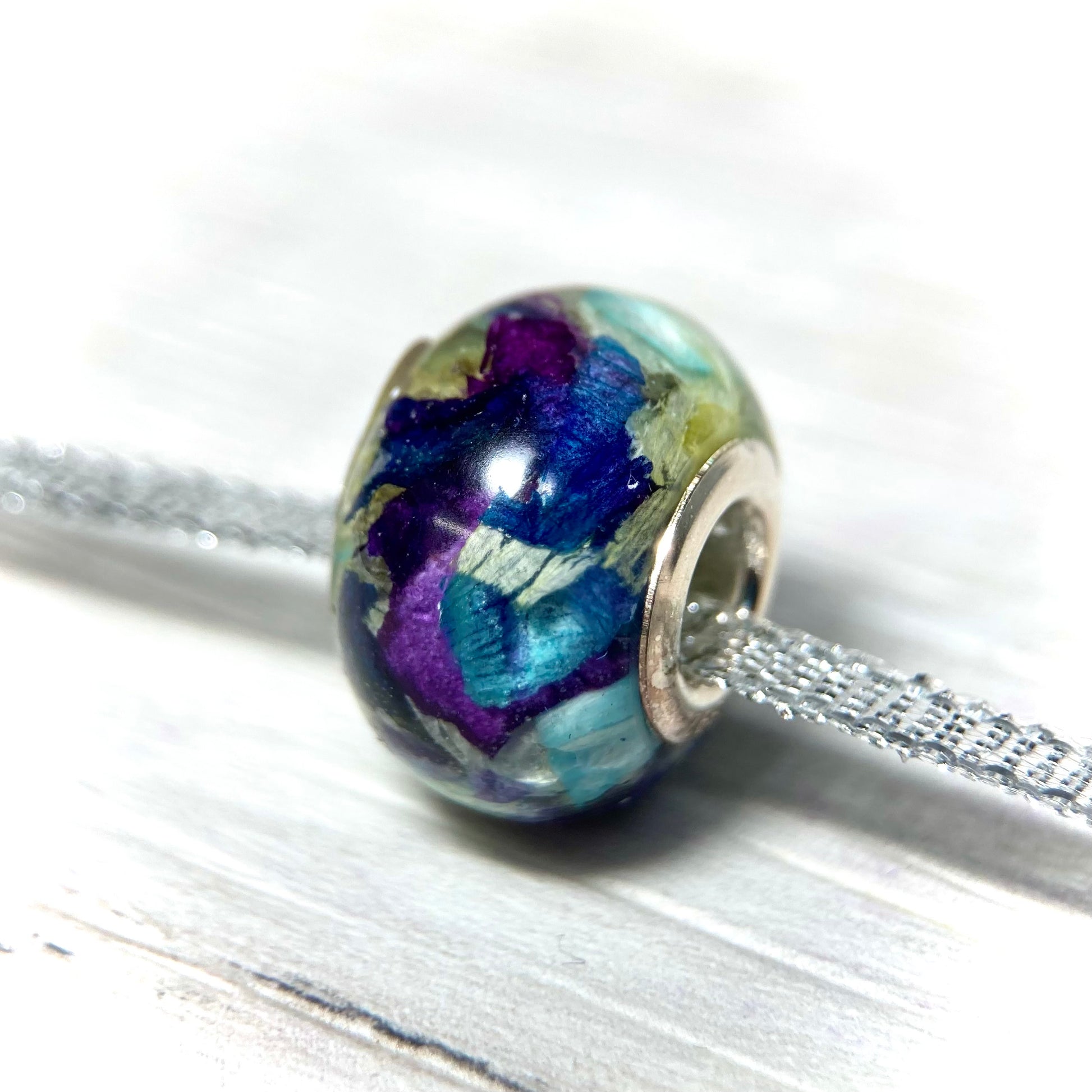 A stunning charm bracelet bead featuring preserved wedding flower petals encased in clear resin with a gleaming sterling silver core. The petals, captured forever in time, serve as a cherished memento of a special day and symbolize the beauty and love of the occasion. A perfect keepsake to wear every day and remember the joy of the wedding day.