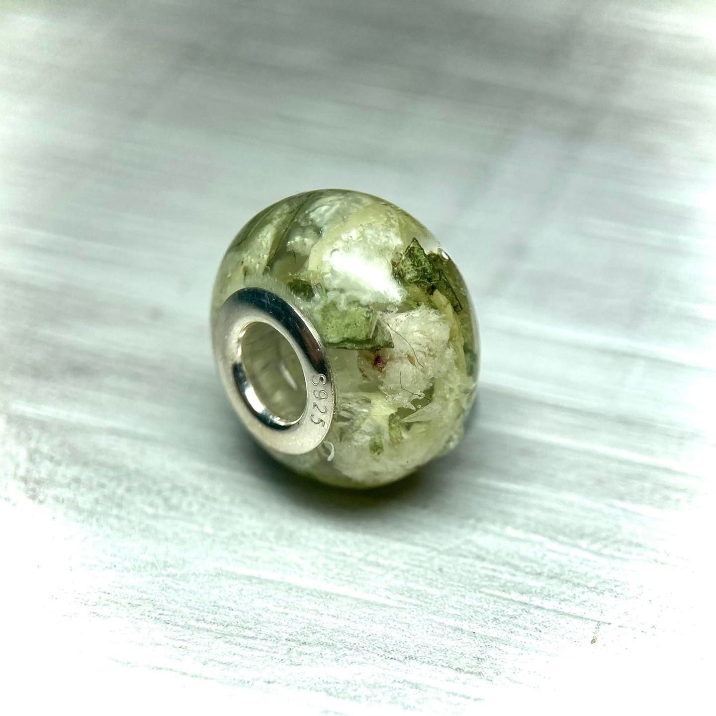 A stunning charm bracelet bead featuring preserved wedding flower petals encased in clear resin with a gleaming sterling silver core. The petals, captured forever in time, serve as a cherished memento of a special day and symbolize the beauty and love of the occasion. A perfect keepsake to wear every day and remember the joy of the wedding day.