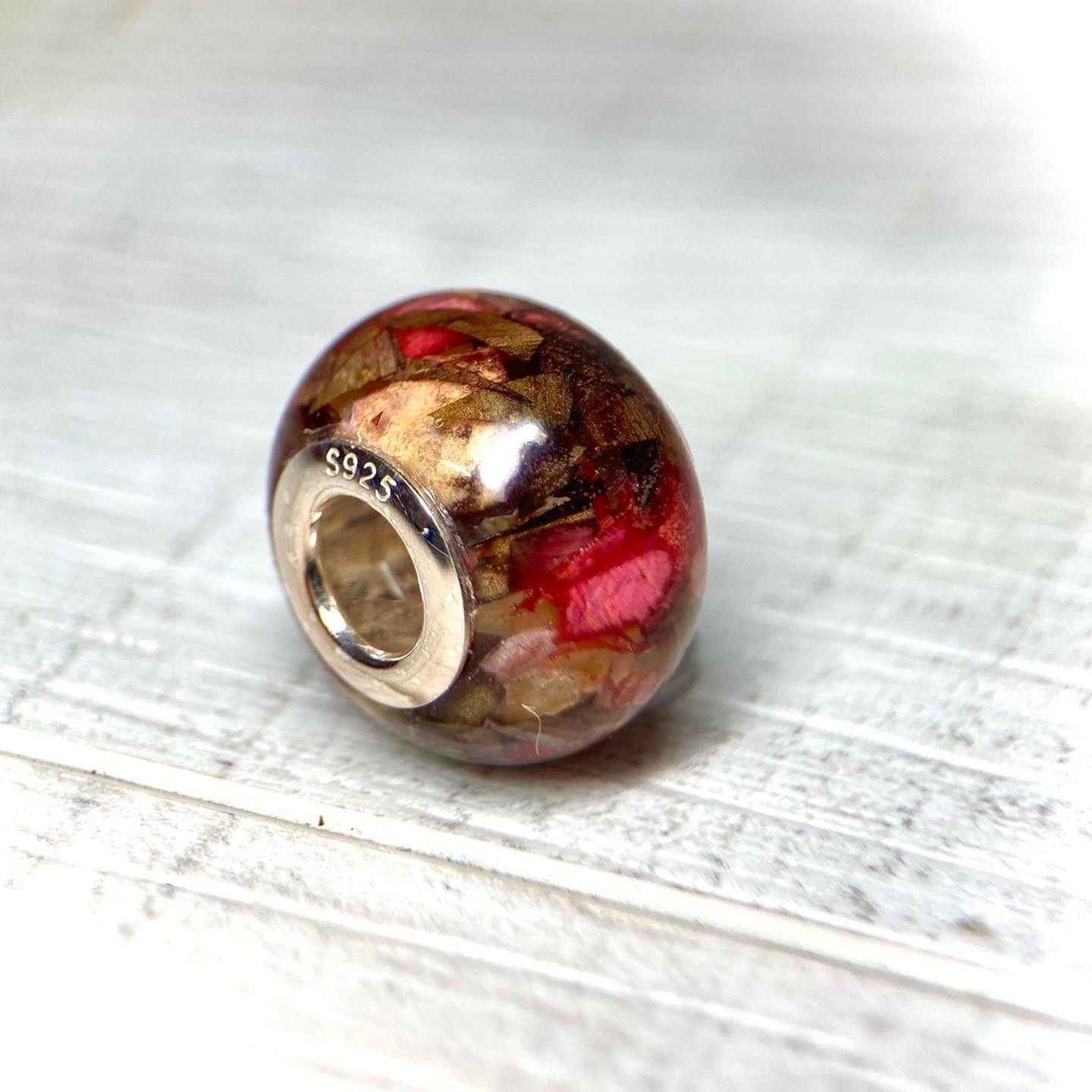 A stunning charm bracelet bead featuring preserved wedding flower petals encased in clear resin with a gleaming sterling silver core. The petals, captured forever in time, serve as a cherished memento of a special day and symbolize the beauty and love of the occasion. A perfect keepsake to wear every day and remember the joy of the wedding day.