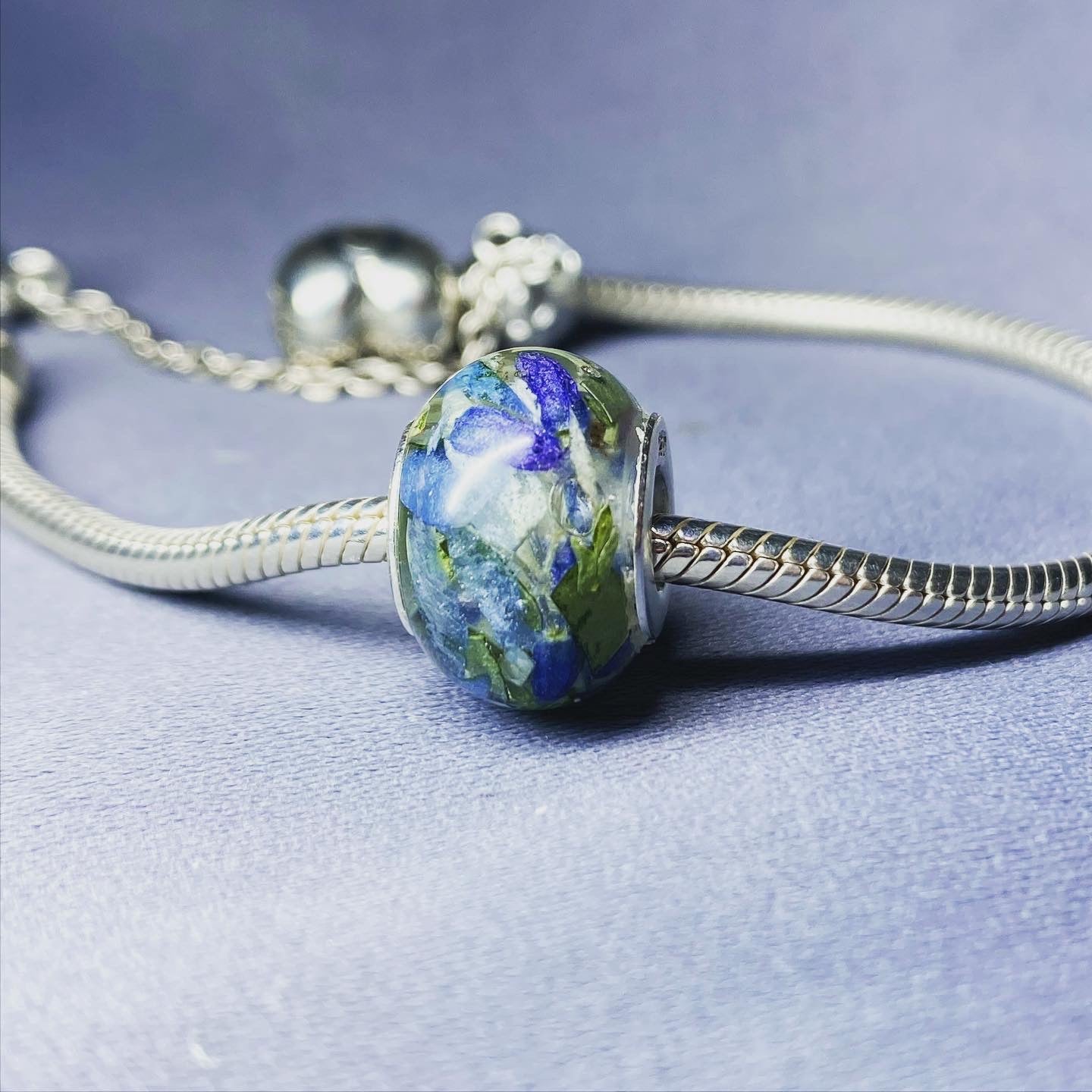 A stunning charm bracelet bead featuring preserved wedding flower petals encased in clear resin with a gleaming sterling silver core. The petals, captured forever in time, serve as a cherished memento of a special day and symbolize the beauty and love of the occasion. A perfect keepsake to wear every day and remember the joy of the wedding day.