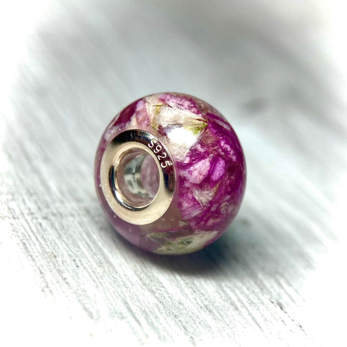 A stunning charm bracelet bead featuring preserved wedding flower petals encased in clear resin with a gleaming sterling silver core. The petals, captured forever in time, serve as a cherished memento of a special day and symbolize the beauty and love of the occasion. A perfect keepsake to wear every day and remember the joy of the wedding day.