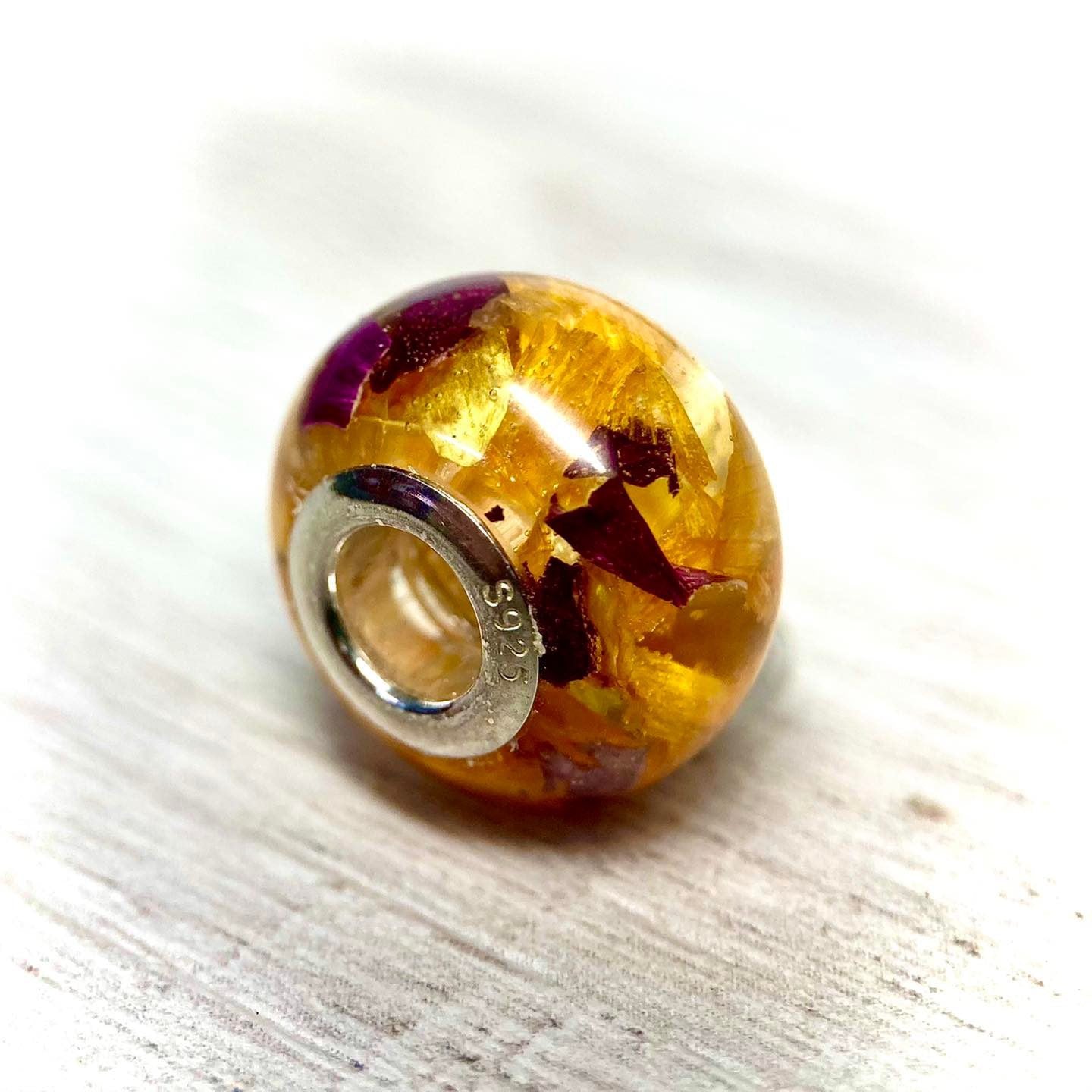 A stunning charm bracelet bead featuring preserved wedding flower petals encased in clear resin with a gleaming sterling silver core. The petals, captured forever in time, serve as a cherished memento of a special day and symbolize the beauty and love of the occasion. A perfect keepsake to wear every day and remember the joy of the wedding day.